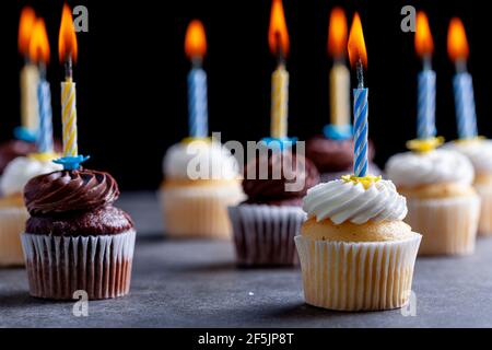 Close up side view image of an assortment of chocolate and vanilla cupcakes with white and brown frostings on. Each have a candle lit against dark bac Stock Photo