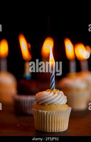 Close up side view image of an assortment of chocolate and vanilla cupcakes with white and brown frostings on. Each have a candle lit against dark bac Stock Photo