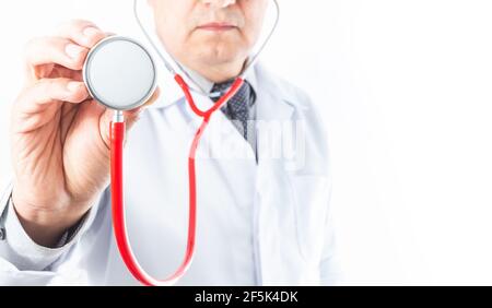 Selective focus of a hand holding a red stethoscope, with a doctor in a white coat and tie out of focus on white background. Good copyspace Stock Photo