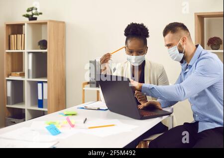 Multiracial colleagues in medical masks work on a laptop discussing a business project. Stock Photo