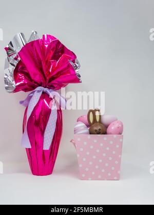 Giant chocolate Easter egg wrapped in pink silver foil next to a pink basket with decorative eggs and a hidden chocolate rabbit Stock Photo