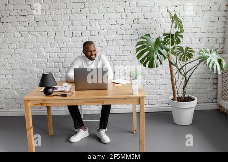 Joyful business male is sitting at the desk in home office, smiling, enjoy job results. Successful young adult man is using laptop, working on project Stock Photo