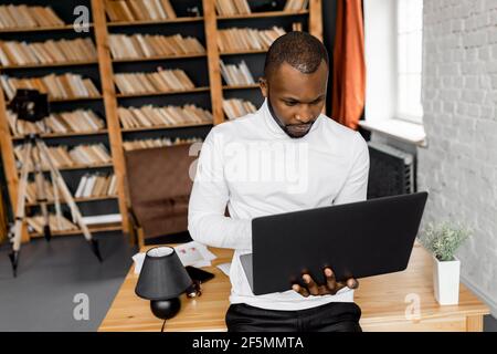 Focused african american businessman is standing in home office, using laptop. Serious male freelancer or student searching ideas for project or Stock Photo