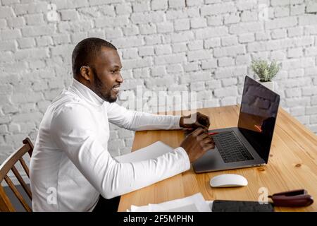Successful male freelancer is using laptop, chatting with coworkers, smiling. Happy young businessman sitting at the desk, discussing ideas for the Stock Photo