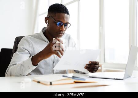 Afro american serious man in earphones drinking coffee while working with papers and laptop in office Stock Photo