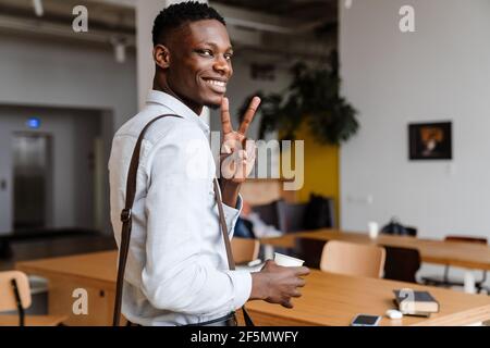 Afro american pleased man gesturing peace sign and drinking coffee while standing indoors Stock Photo