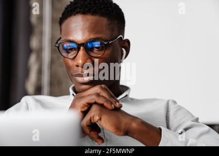 Afro american focused young man in eyeglasses working with laptop indoors Stock Photo