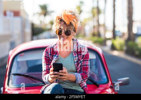 Trendy female young adult caucasian woman use modern phone connection outdoor near her pretty red car in travel and independent lifestyle - concept of Stock Photo