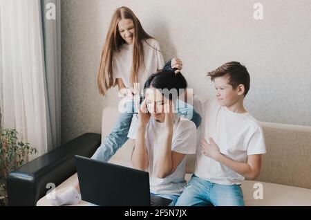 An irritated mother sits on the couch and works at home on a laptop, active children play with their mother's hair, do her hair. Tired, stressed mom Stock Photo