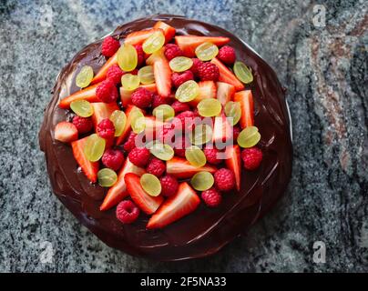 Top-Down Dark Chocolate Cake with Fruit on Marble Kitchen Counter. Tart with Chocolate Icing and Strawberries, Grapes and Raspberries. Stock Photo