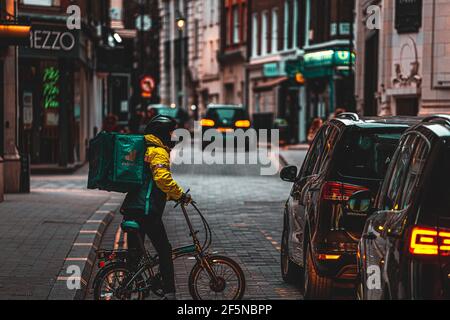 London UK February 2021 Lone Deliveroo food delivery man trying to make his way on a bicycle through a traffic jam, pushing his bike in between the ca Stock Photo
