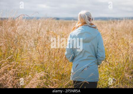A young blonde female caucasian alone in nature standing in long grass in the rural English countrysie looking out in contemplation. Stock Photo