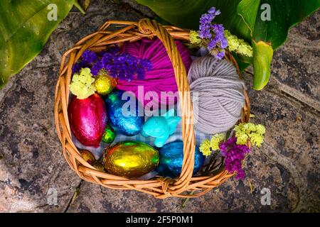 A basket decorated for an egg hunt. It has eggs wrapped in color tin foil, a toy of a rabbit and some balls of yarn. It's surrounded by vegetation, cl Stock Photo