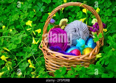 A basket decorated for an egg hunt. It has eggs wrapped in color tin foil, a toy of a rabbit and some balls of yarn. It's surrounded by vegetation, cl Stock Photo