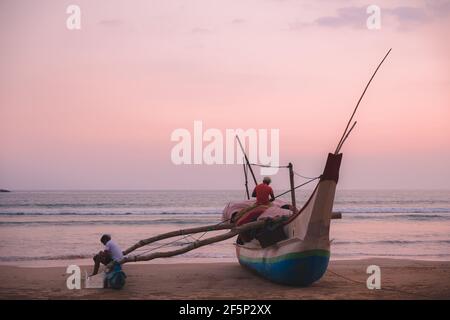 Local Sri Lankan fisherman at sunrise or sunset prepare for morning or evening fishing on a traditional catamaran boat from ashore on Weligama Beach, Stock Photo