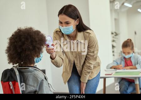 Beautiful young female teacher wearing protective mask measuring temperature, screening little school boy with digital thermometer Stock Photo