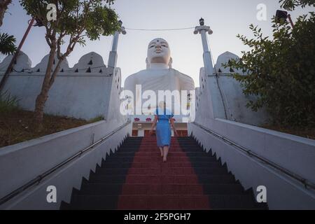 A young female caucasian tourist ascends the steps to the giant white Buddha statue at Sri Maha Bodhi Viharaya, a Buddhist temple at Bahirawakanda in Stock Photo