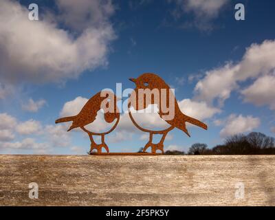 A decorative pair of robins laser cut in rusting steel mounted on a wooden base with light cloud and blue sky behind Stock Photo