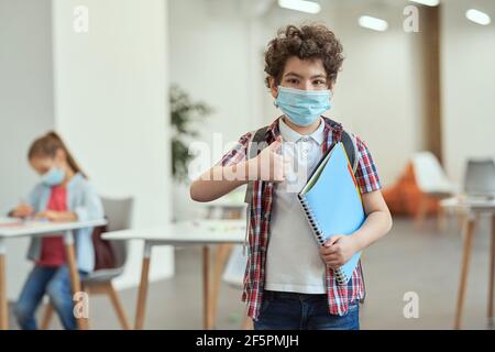 Portrait of little school boy wearing mask to prevent the spread of Covid19 looking at camera, showing thumbs up, holding textbook while posing in a Stock Photo