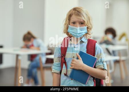 Portrait of calm little school boy wearing mask to prevent the spread of Covid19 looking at camera, holding notebook while posing in a classroom Stock Photo