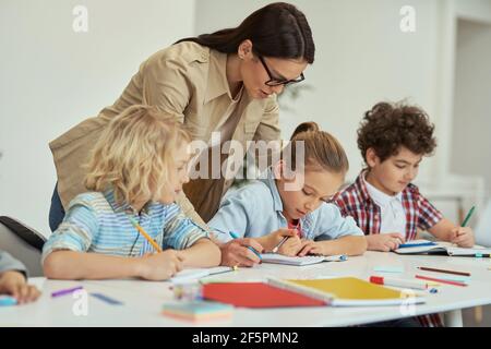 Excellence. Kind young female teacher in glasses helping her little schoolchildren in a classroom. Kids sitting at the table, studying in elementary Stock Photo