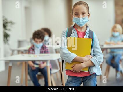 Portrait of happy little school girl wearing mask to prevent the spread of Covid19 looking at camera, holding textbooks while posing in a classroom Stock Photo