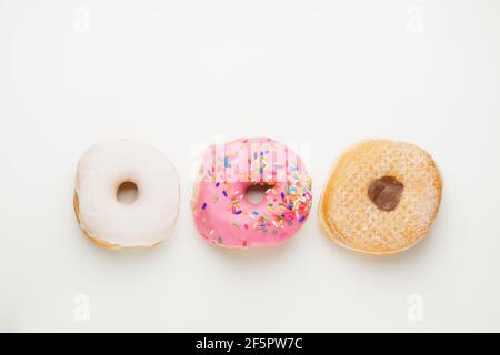 Overhead flat lay view of three fresh, colourful ring doughnuts in a row isolated on a plain white surface and background. Stock Photo