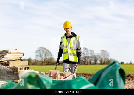 A young adult male builder wearing a high visibility vest and hard hat pushing a wheelbarrow full of bricks while on a building site Stock Photo