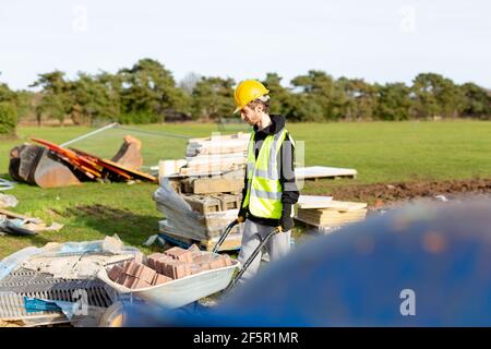 A young adult male builder wearing a high visibility vest and hard hat pushing a wheelbarrow full of bricks while on a building site Stock Photo