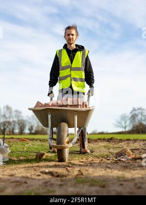 A young adult male builder wearing a high visibility vest and hard hat pushing a wheelbarrow full of bricks while on a building site Stock Photo