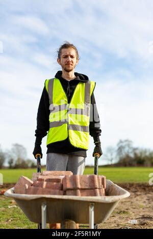 A young adult male builder wearing a high visibility vest and hard hat pushing a wheelbarrow full of bricks while on a building site Stock Photo