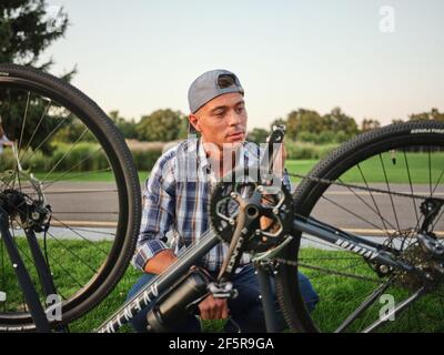 Breakage. Caucasian young man looking concentrated while checking pedals on his bike, kneeling on grass in public park Stock Photo