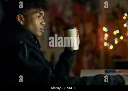 Need some sleep. Thoughtful young guy with piercing holding disposable cup of coffee, sitting at the table and working on laptop from home late at Stock Photo