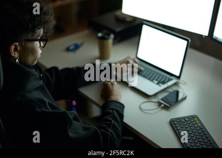 Young guy wearing glasses sitting at the table in front of many computer monitors and using laptop while working late at night Stock Photo