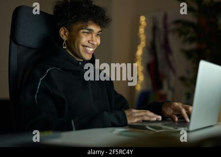 Cheerful young guy with piercing smiling while sitting in front of laptop screen late in the evening Stock Photo