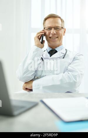 Cheerful male doctor talking on mobile phone at work Stock Photo