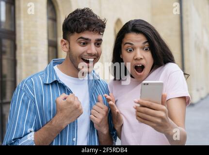 Emotional couple wins online lottery, celebration success. Excited friends using smartphone, shopping online Stock Photo
