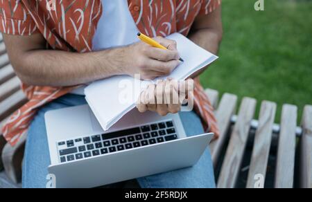 Close up of male hands taking notes on notebook, using laptop computer, planning project. Business man working online outdoors, focus on hand Stock Photo