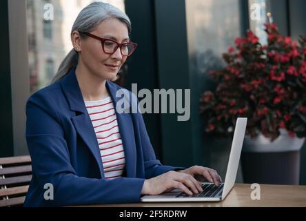 Mature businesswoman using laptop computer, working online. Portrait of middle aged asian copywriter wearing stylish eyeglasses typing on keyboard Stock Photo