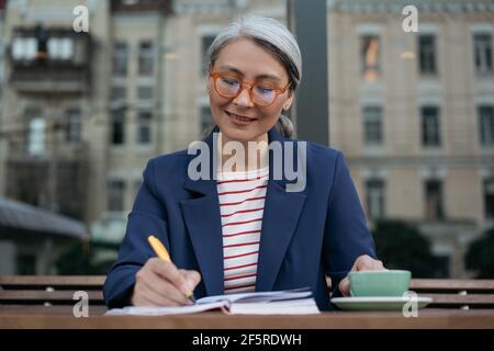 Portrait of middle aged asian writer taking notes. Mature businesswoman wearing stylish eyeglasses working project, planning startup Stock Photo