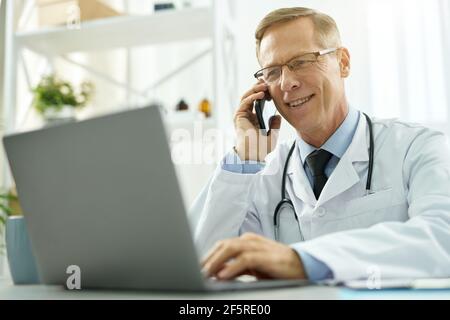 Joyful doctor talking on cellphone and using notebook in clinic Stock Photo