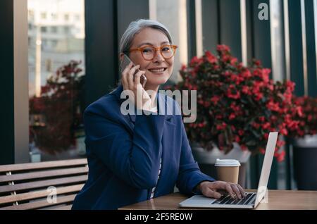 Mature asian woman wearing stylish eyeglasses talking on mobile phone, sitting at workplace. Portrait of middle aged businesswoman using laptop Stock Photo
