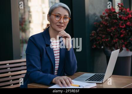 Pensive mature business woman looking at camera, sitting at workplace. Beautiful middle aged freelancer working, using laptop, planning project