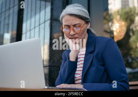 Tired asian businesswoman working project, using laptop, searching information. Portrait of frustrated mature woman sitting at workplace