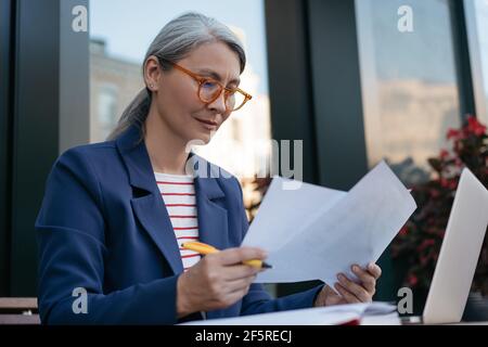 Pensive mature businesswoman reading contract, planning project. Portrait of middle aged asian secretary reading, working with documents Stock Photo