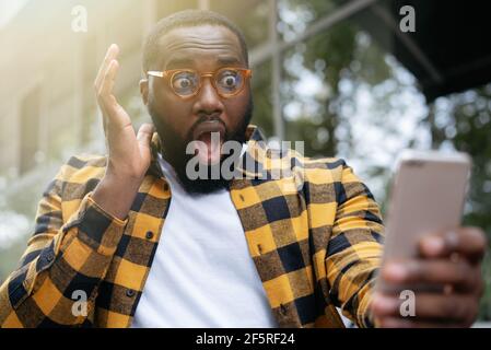 Shocked man with open mouth using mobile phone, looking at digital screen. Portrait of emotional African American freelancer missed deadline Stock Photo