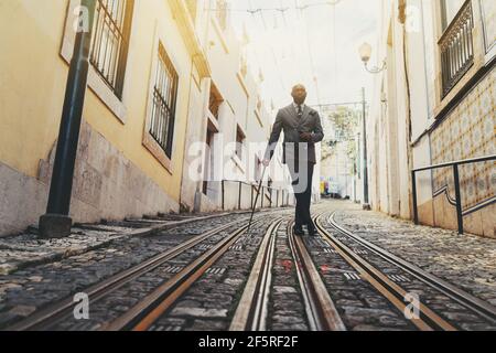 A dapper black senior man with a well-groomed beard and totally hairless is leaning against his fancy walking stick while standing on the tramway trac Stock Photo