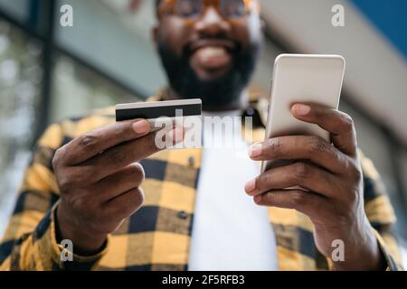 Close up of man hands holding credit card and mobile phone, shopping online. Happy African American freelancer receive payment, focus on hands Stock Photo