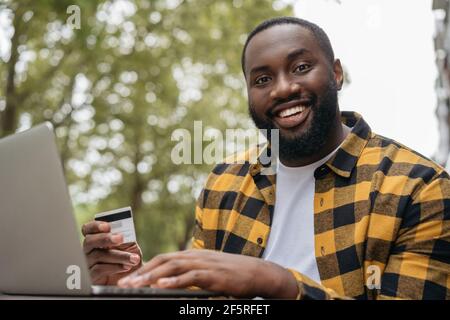 Portrait of young smiling African American man holding credit card, shopping online. Black Friday concept. Happy successful freelancer receive payment Stock Photo