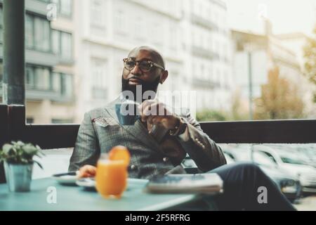 The portrait of a handsome stylish wealthy African guy with a beautiful black beard, in glasses, bald, in an elegant suit, sitting on a rainy morning Stock Photo